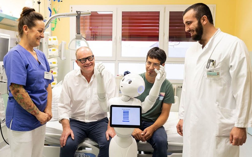 Vier Menschen und ein humanoider Roboter sind um ein Klinikbett versammelt