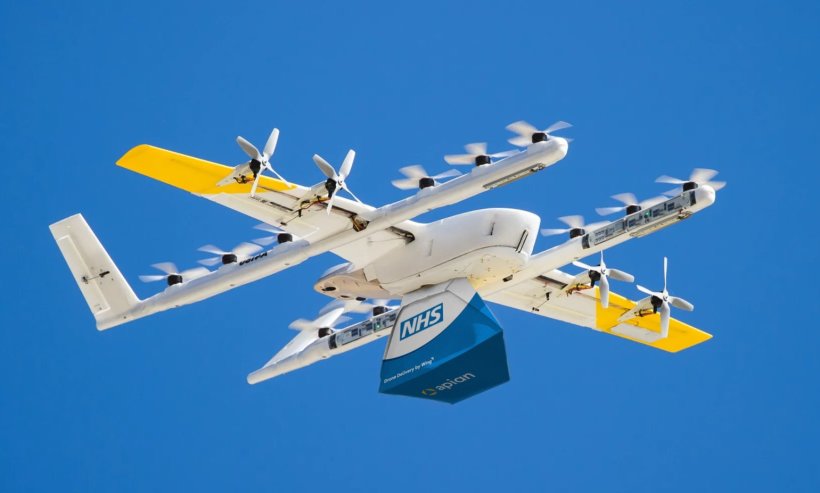 A white drone with yellow wingtips carrying a blue box labelled NHS in the...