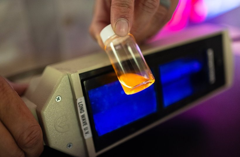 A fluorescent orange liquid in a glass vial is held in front of a blue glowing...