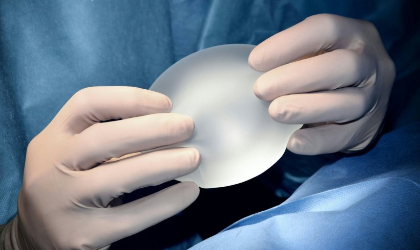 A silicone breast implant is held by a surgeon in white rubber gloves