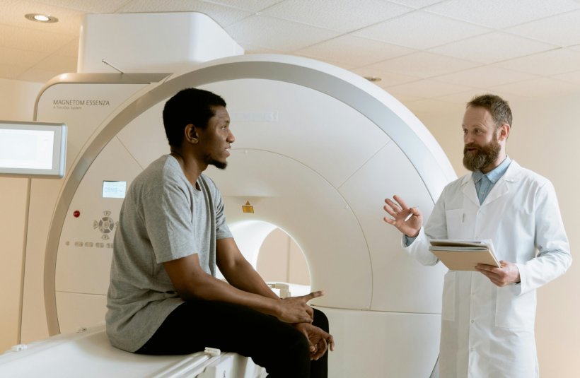 A clinician in a white doctors coat talks to his patient, who is sitting on the...