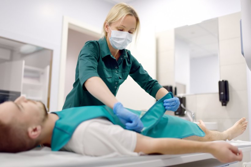 A radiologist wearing a protective mask puts a lead apron on a lying patient