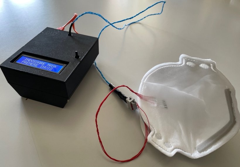 A black box with a blue electronic display is connected to an FFP2 face mask...