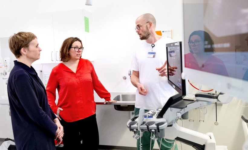Three people are standing in a hospital room, facing each other. The man on the...