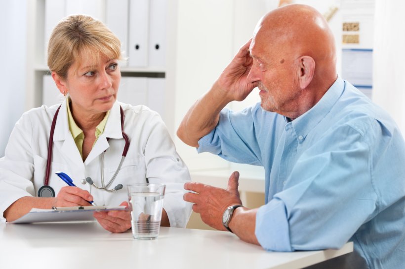 A doctor holding a clipboard is talking to her elderly patient, sitting at the...