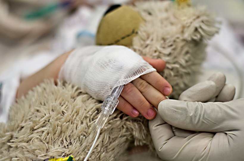 Die bandagierte Hand eines Kindes hält einen Teddy. Die Hand eines Mediziners...