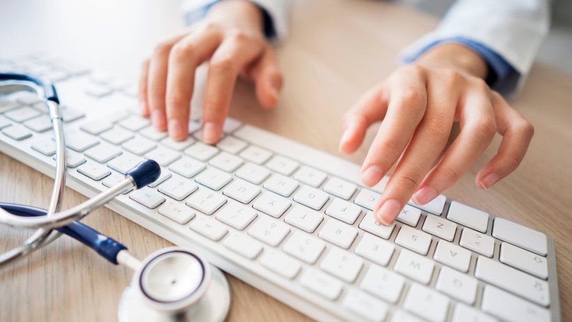 Die Hände einer Ärztin bedienen eine weiße Computer-Tastatur. Am oberen Rand...