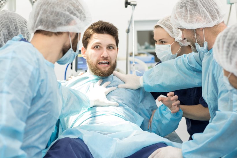 A scared-looking man is sitting upright on a surgery table, surrounded by...