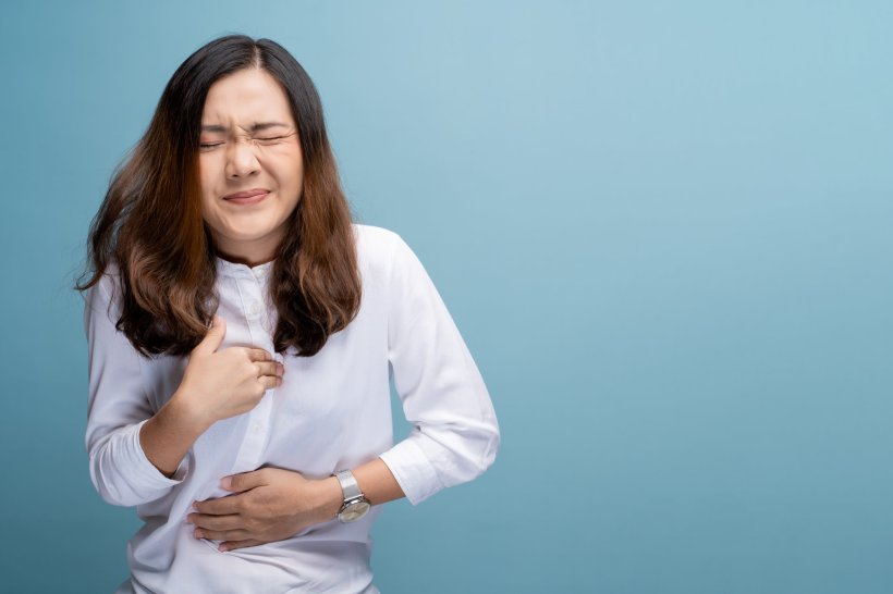 A woman in a white shirt holding her chest and stomach in pain, against a blue...