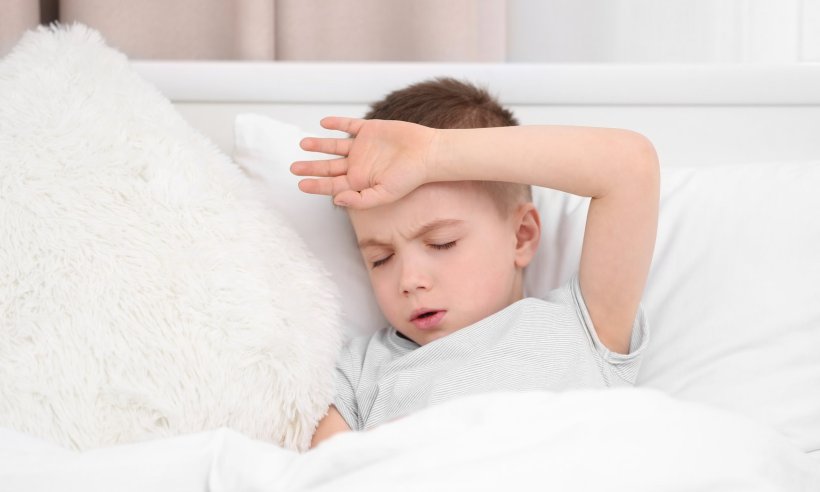 Ein kleiner Junge liegt zuhause im Bett und hält sich mit der linken Hand die...