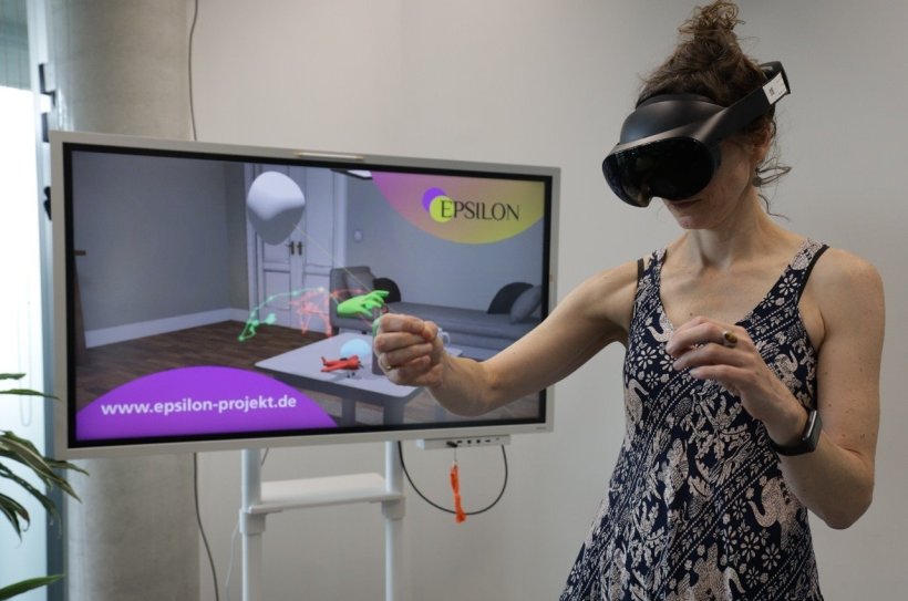 Eine Frau mit VR-Headset und VR-Controllern steht vor einem großen Bildschirm,...