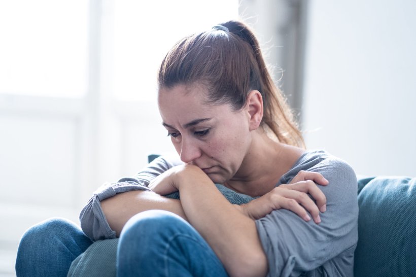 A sad looking woman is sitting on her sofa at home with her knees pulled to her...