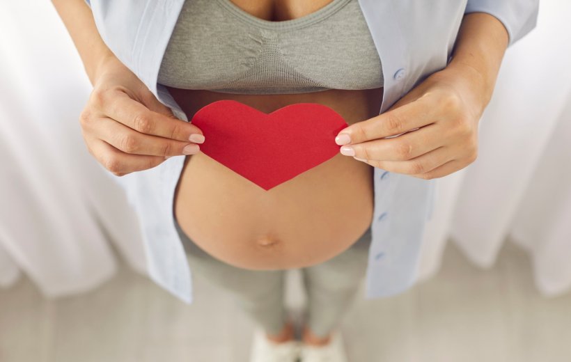 Eine schwangere Frau hält mit beiden Händen ein rotes Herz aus Papier vor...