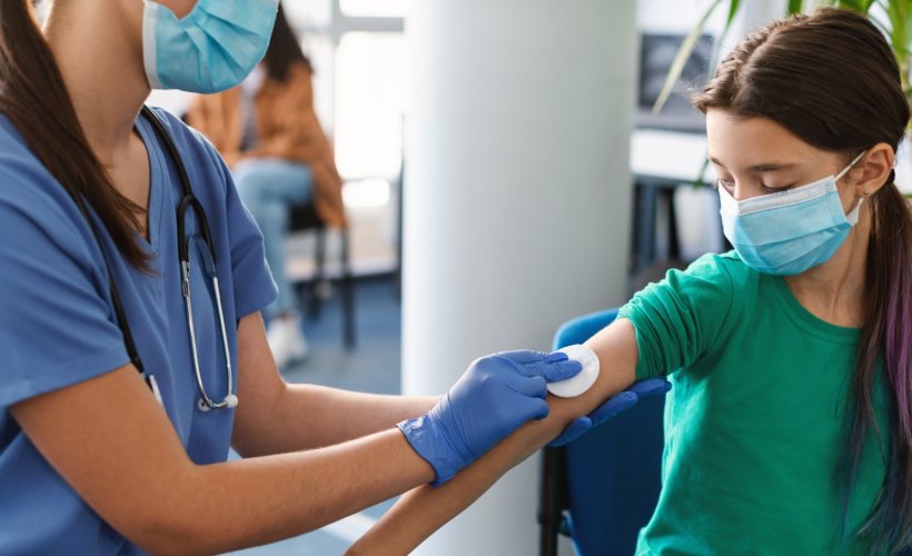 A young girl wearing a face mask receives a vacine shot from a healthcare...