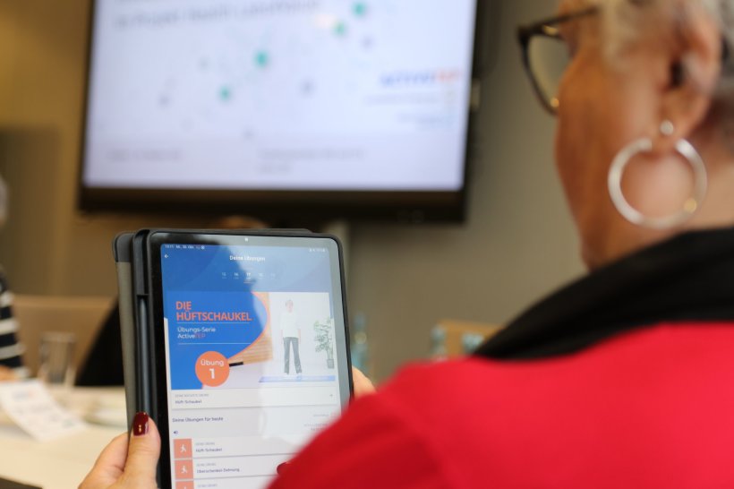 Eine ältere Frau hält ein Tablet in der Hand, auf dem eine Übung für die...