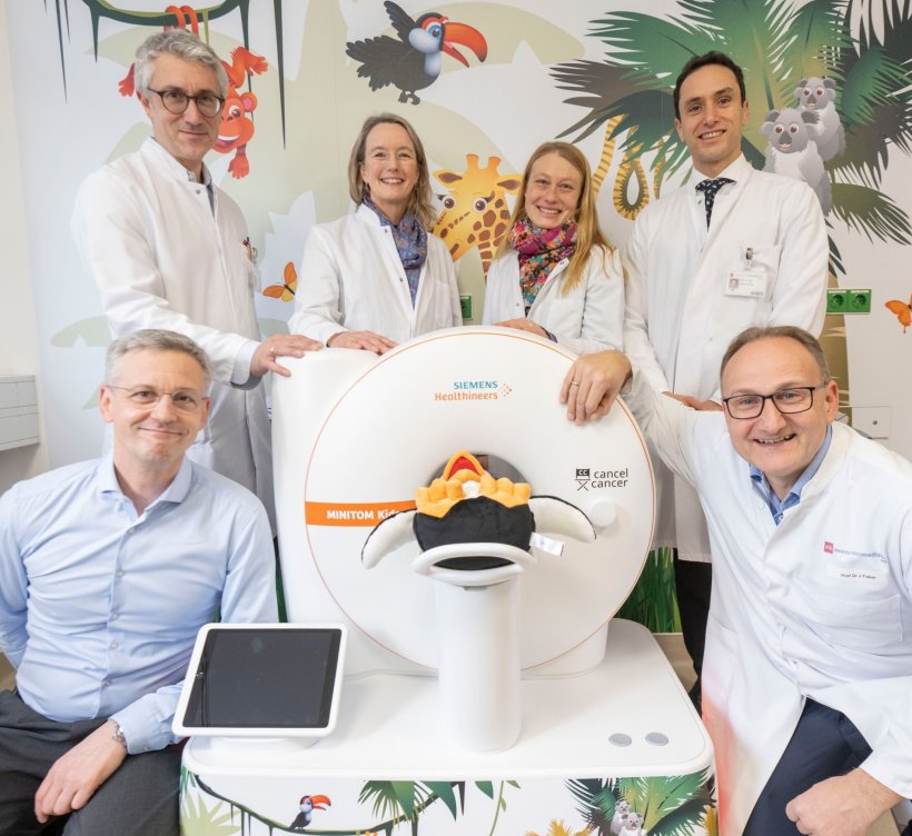 Sechs Personen stehen und sitzen um ein Miniatur-Modell eines MRT und blicken...