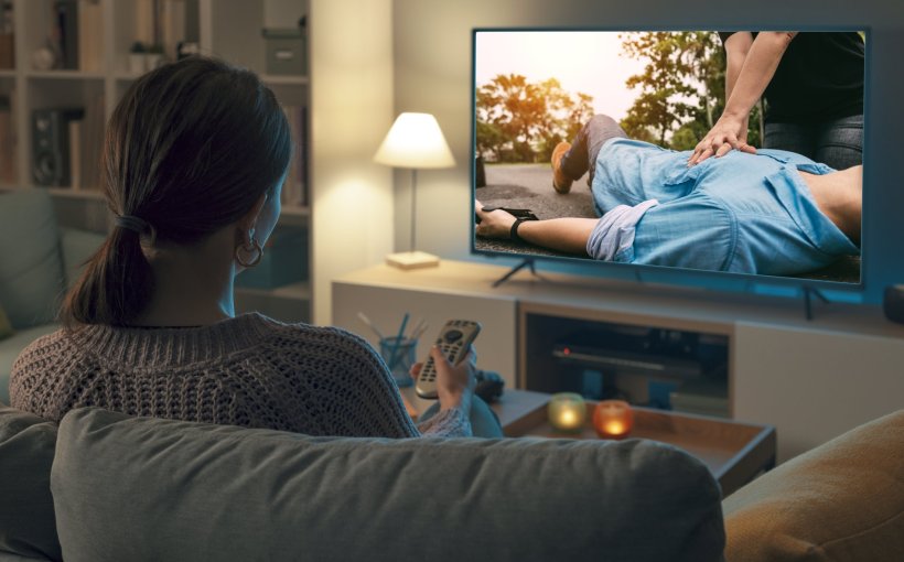 A woman is watching TV on her sofa, with the remote control pointed at the TV....