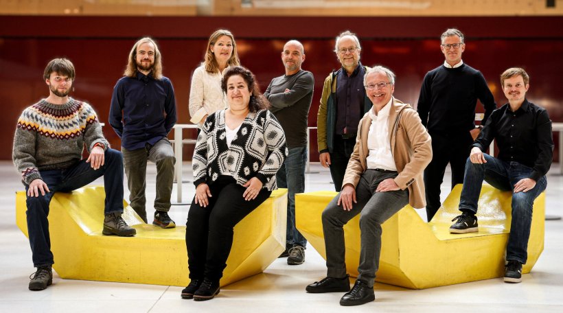 Eine Gruppe aus neun Personen sitzt für ein Gruppenbild in einer großen Halle...