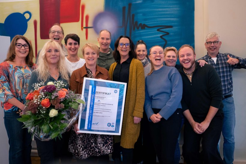 Zwölf Personen stehen zusammen für ein Gruppenfoto vor einer bunten Wand....