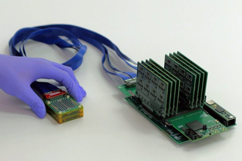 A hand in a blue rubber glove holds a small green box, whcih is connected via a...