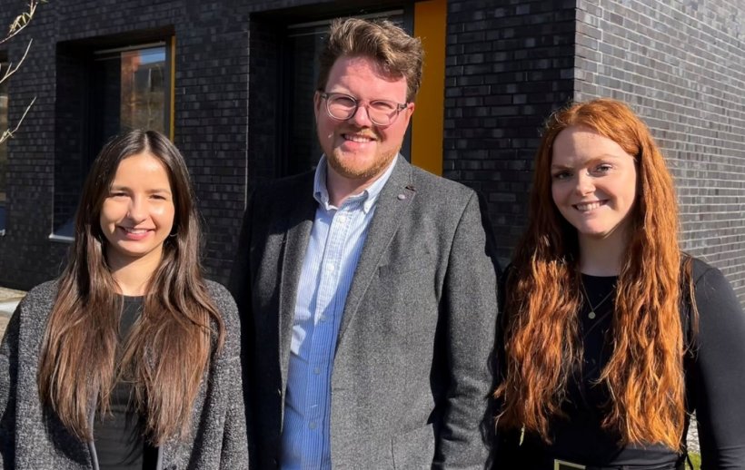 Drei Personen stehen für ein Teamfoto nebeneinander. Im Hintergrund ist ein...