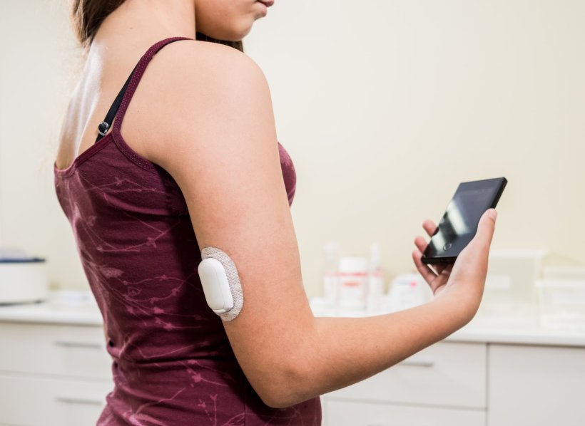 Eine junge Frau mit einem CGM-Pflaster auf dem linken Oberarm schaut auf ihr...