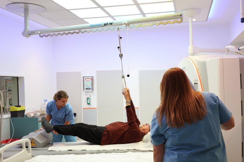 Two medical professionals are helping a patient stand up from a hospital bed,...