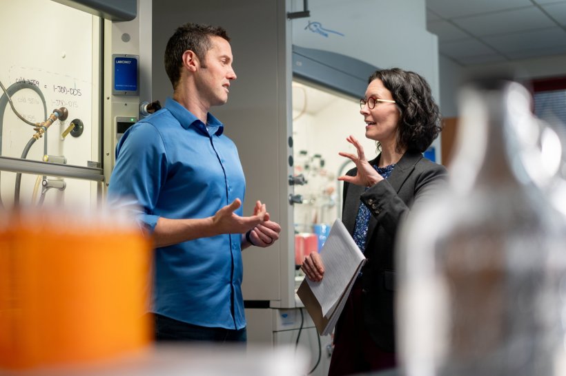 Two persons are standing in a laboratory, facing each other. In the foreground,...