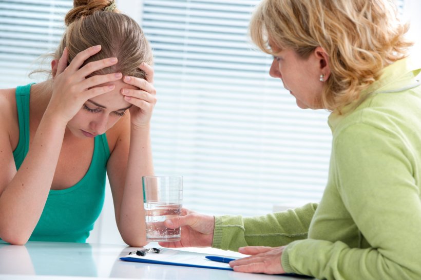 A stressed out woman is sitting at a doctors office desk with her head buried...