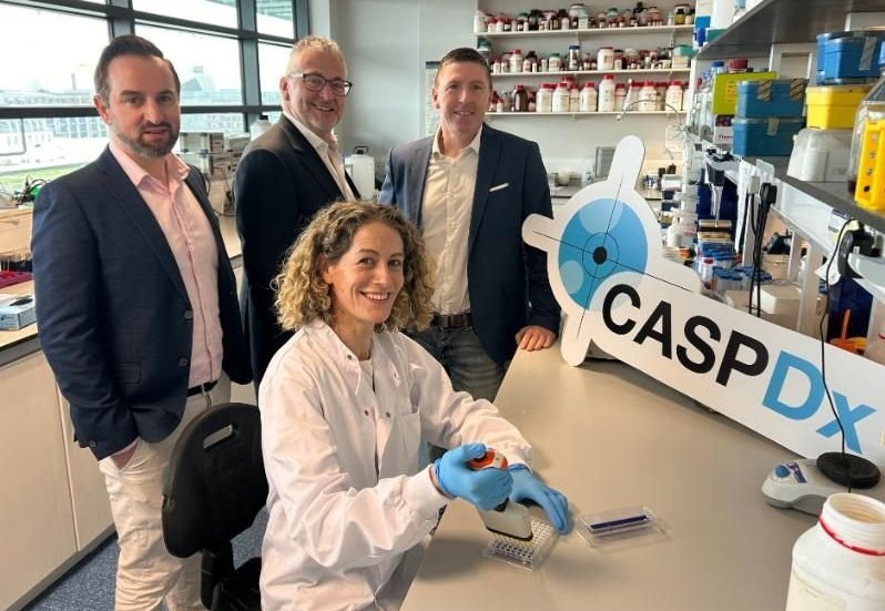 Three men are standing around a woman sitting at a laboratory desk, operating a...