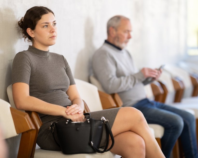 A young woman is sitting in a chair of a hospital waiting room, looking...