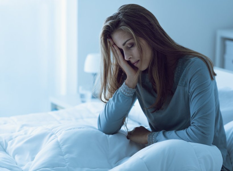 A depressed-looking young woman is sitting in her bed, holding her head with...