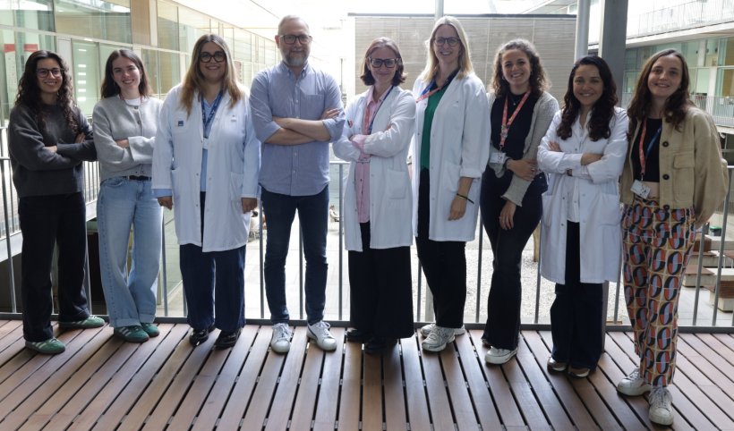 Nine persons are standing in a row for a team photo. In the background, the...