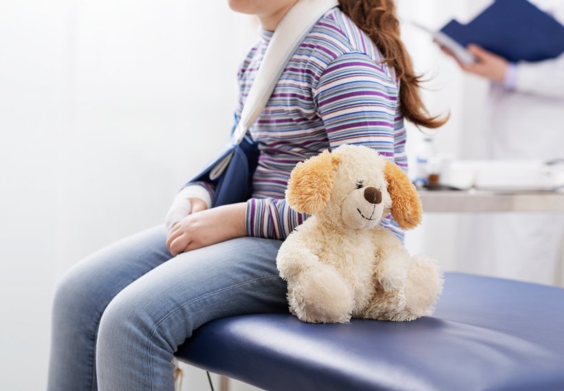 A young girl is sitting at a doctors office with a broken arm with a teddy bear