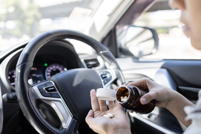 A woman is sitting in the drivers seat of her car, putting pills from a pill...