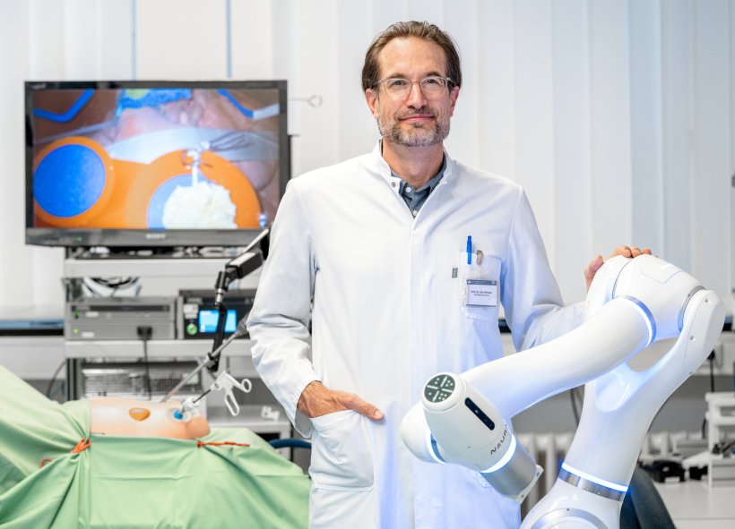 Prof. Dirk Wilhelm steht neben einem weißen Roboterarm. Im Hintergrund ist ein...
