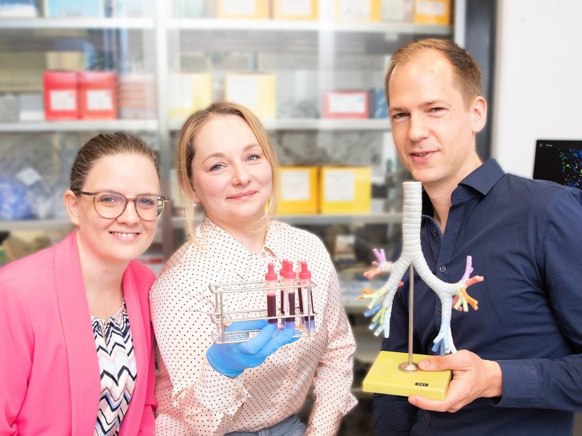 Drei Forscher stehen in einem medizinischen Labor. Die Frau in der Mitte hält...