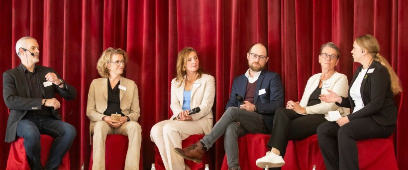 Paneldiskussion mit sechs Personen vor einem dunkelroten Samtvorhang