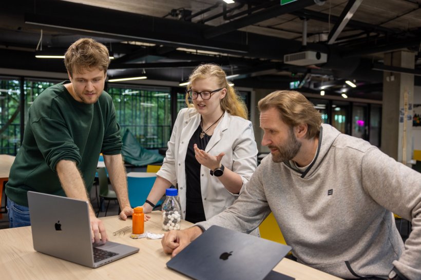 Three researchers are gathered around a desk with two laptop computers, looking...