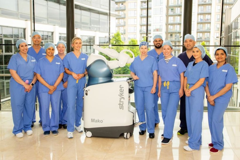 A group photo of eleven surgeons standing around a robotic surgery system in a...