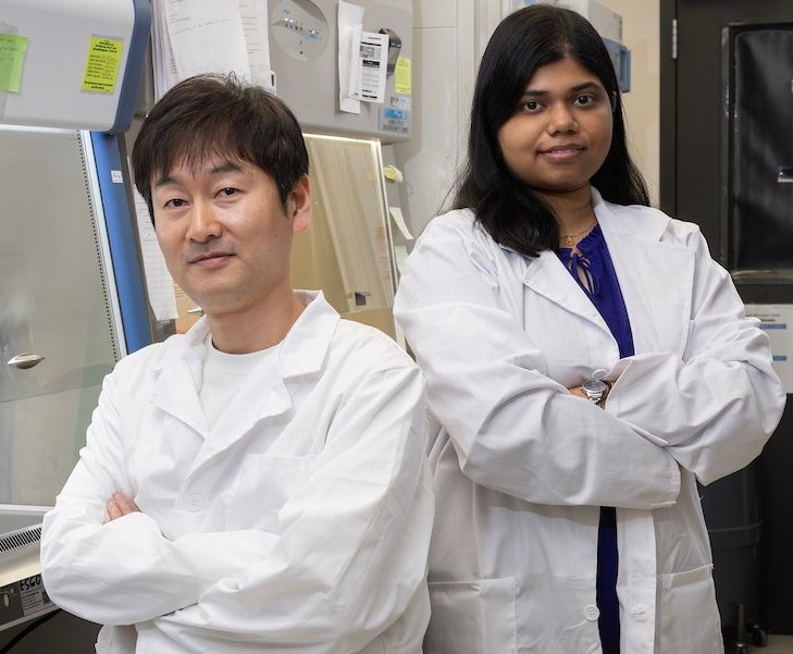 Two researchers wearing white lab coats standing back-to-back with arms crossed...