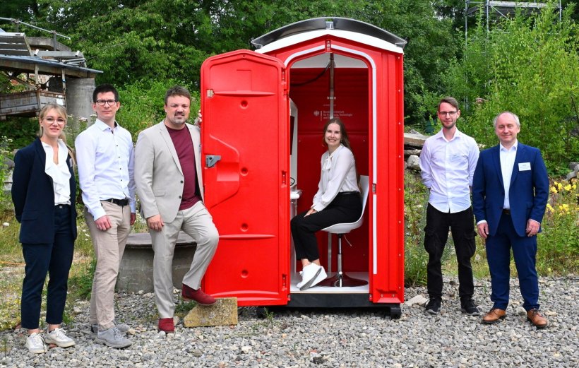 Sechs Personen stehen und sitzen in einem Außengelände um eine rote WC-Kabine