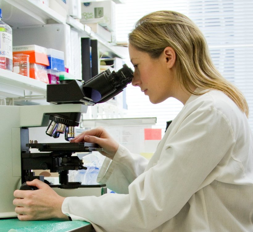 A scientist in a white lab cat is sitting in front of a microscope. She is...