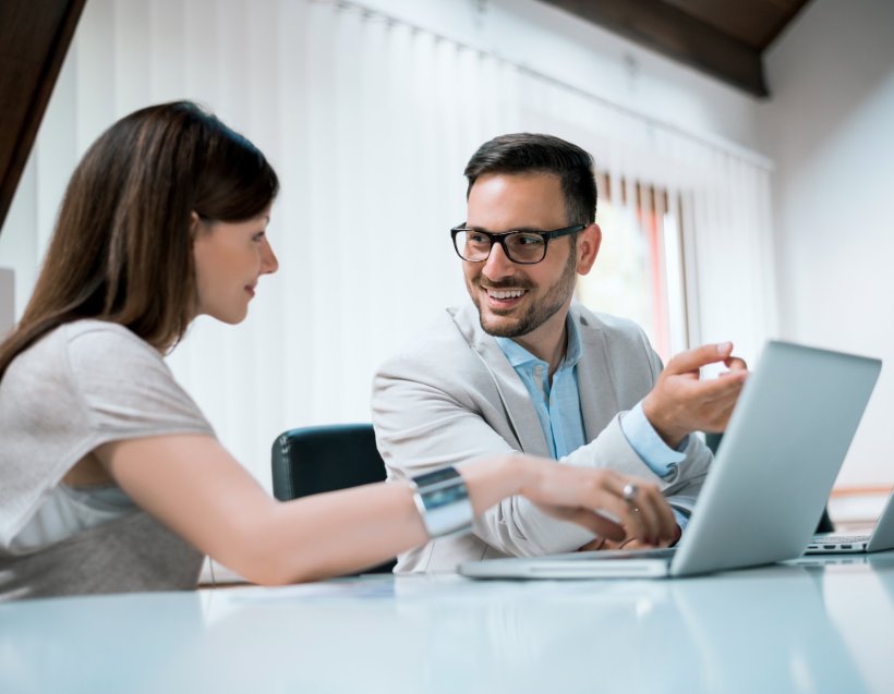 Two smiling young businesspeople are sitting at a desk in front of a laptop
