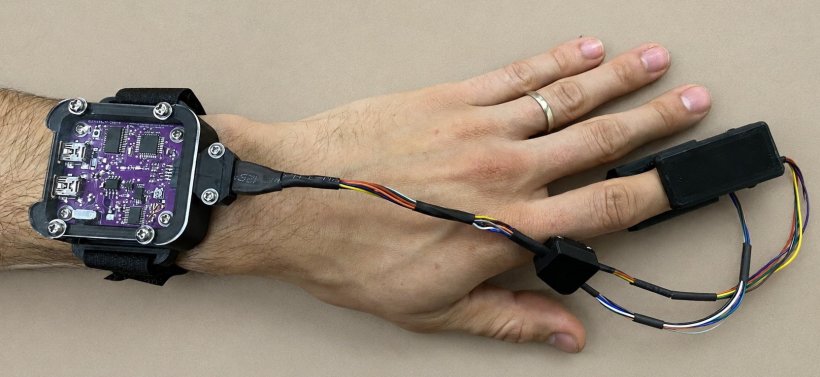 A left hand wearing a prototype version of a pulse oxymeter, with a small...