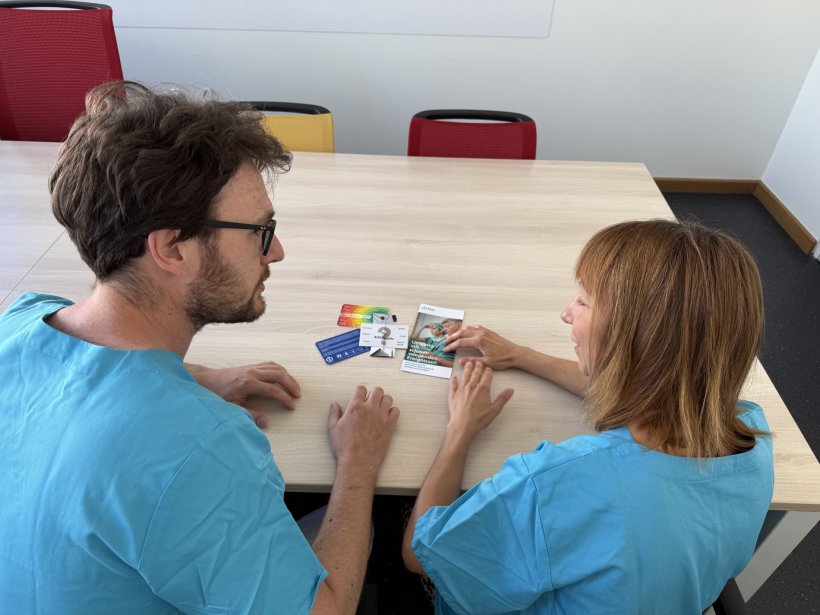 Zwei Klinikmitarbeiter sitzen nebeneinander an einem Holztisch, auf dem mehrere...