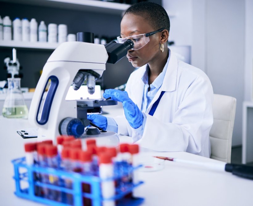 A researcher is sitting in front of a microscope on a white desk. In the...