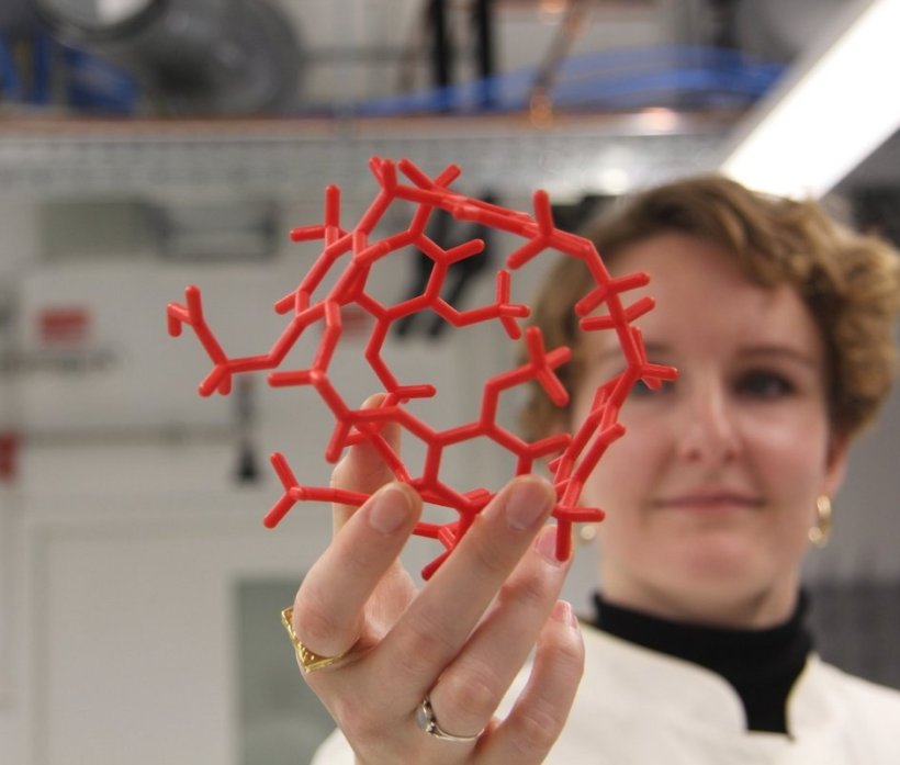 Eine Wissenschaftlerin im weißen Laborkittel hält ein rotes Molekülmodell...