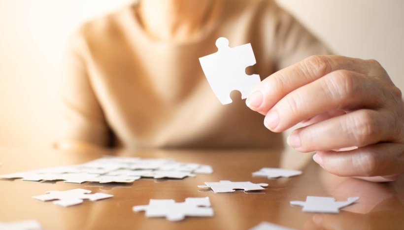 Elderly female hands trying to connect pieces of white jigsaw puzzle on wooden...