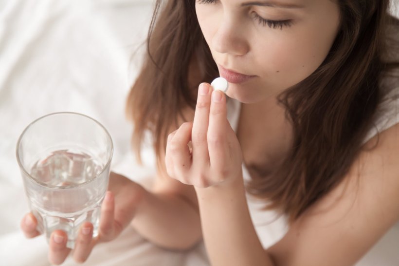 women with headache taking painkiller pill, holding glass of water